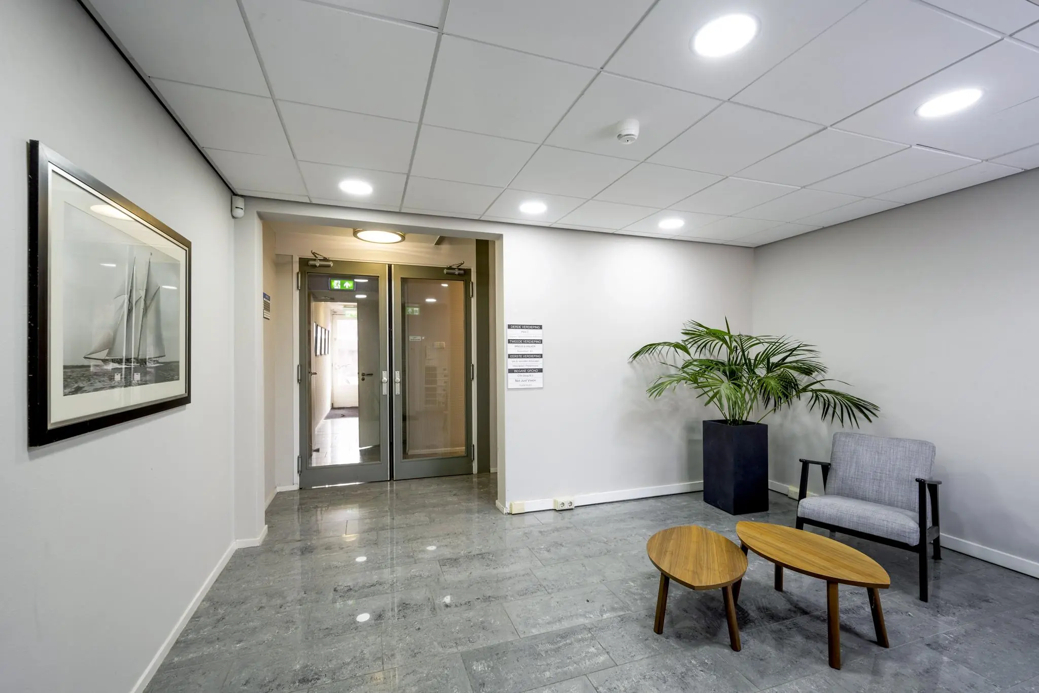 Wachtruimte met grijze vloer, moderne stoelen, plantenbak, kunst aan de muur en glazen deuren naar de gang in gebouw aan de Nieuwe Gracht.